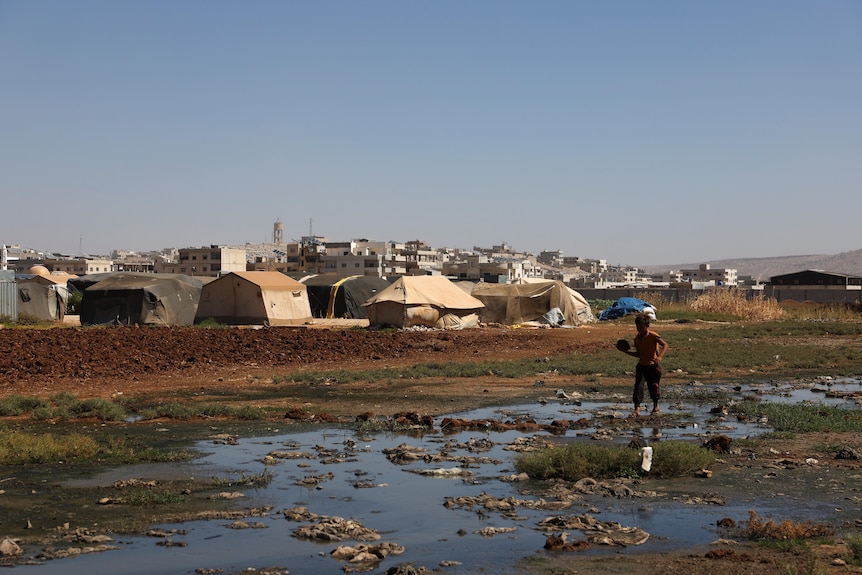 A child walks in a muddy stream next to a tent city. 