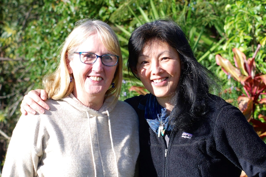 Two smiling women standing beside each other posing for a photo with bushland as background.