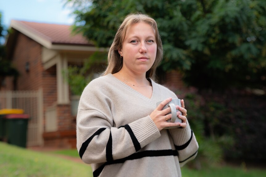 A woman stands in front of a house, holding a coffee cup and looking serious.