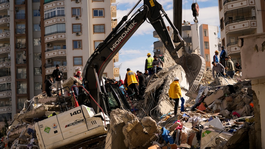 A digger sits on top of rubble as people surround it. 