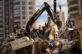 A digger sits on top of rubble as people surround it. 