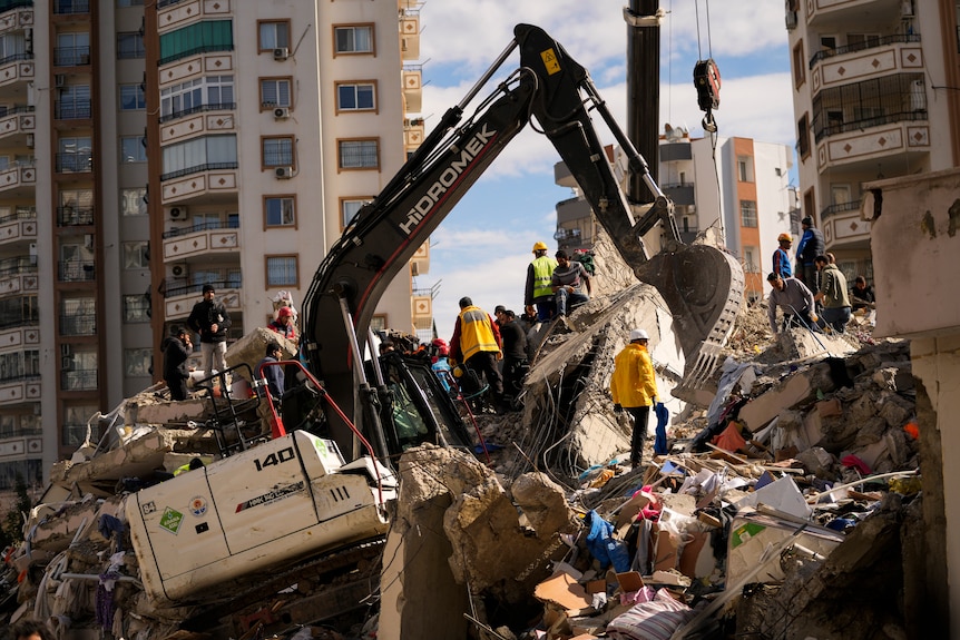 A digger sits on top of rubble as people surround it. 