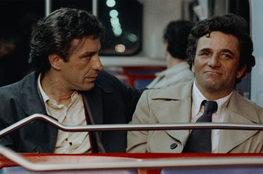 Two middle aged men with dark hair wear button up shirts and trench coats, one with tie, sit and talk on bus at night time.