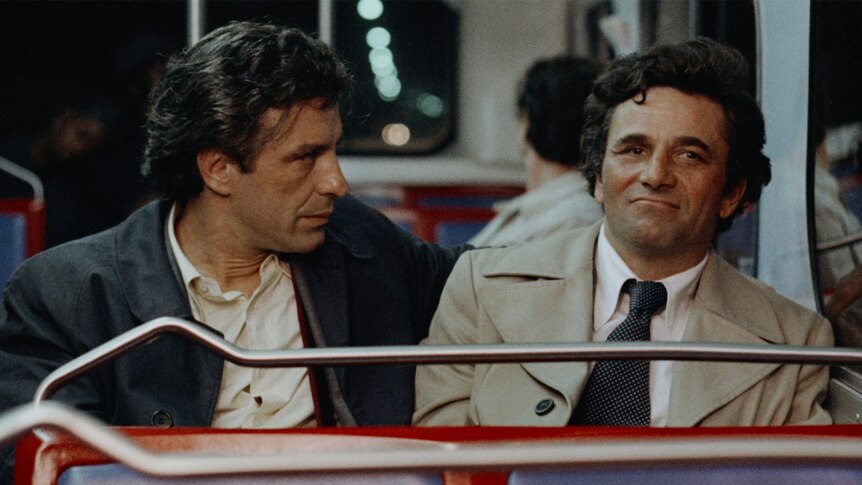 Two middle aged men with dark hair wear button up shirts and trench coats, one with tie, sit and talk on bus at night time.