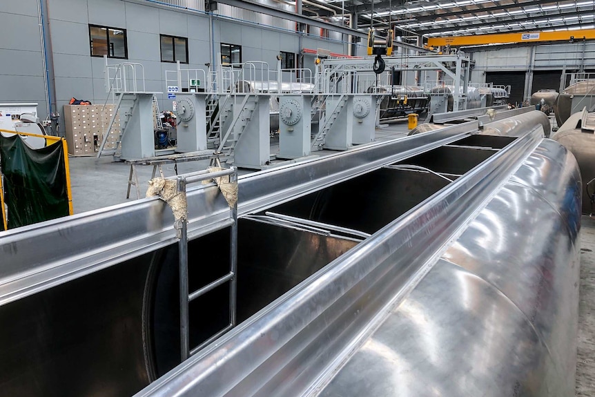 A large silver tank with a ladder leading into it, with a brightly lit silver factory in the background.