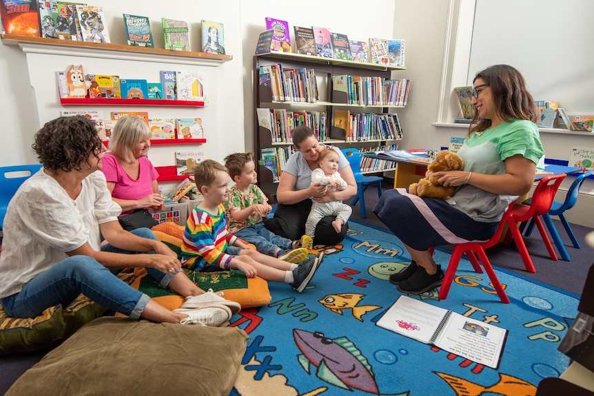 A group of parents and children sit on the floor at a public library listening to a staff member read a book