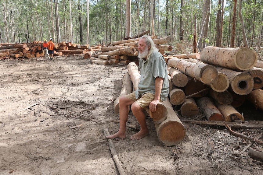 An older man sits barefoot on a pile of logged trees in a forest