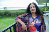 A middle-aged woman with long, dark hair, wearing a colourful top with Indigenous designs stands in a coastal area.