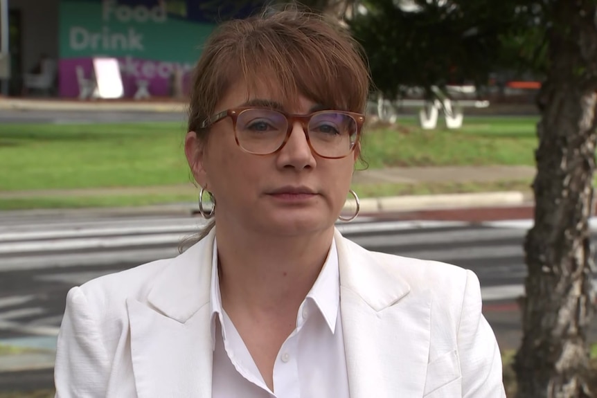 A woman with glasses and a fringe wearing a white shirt and blazer stands in front of a grassed area