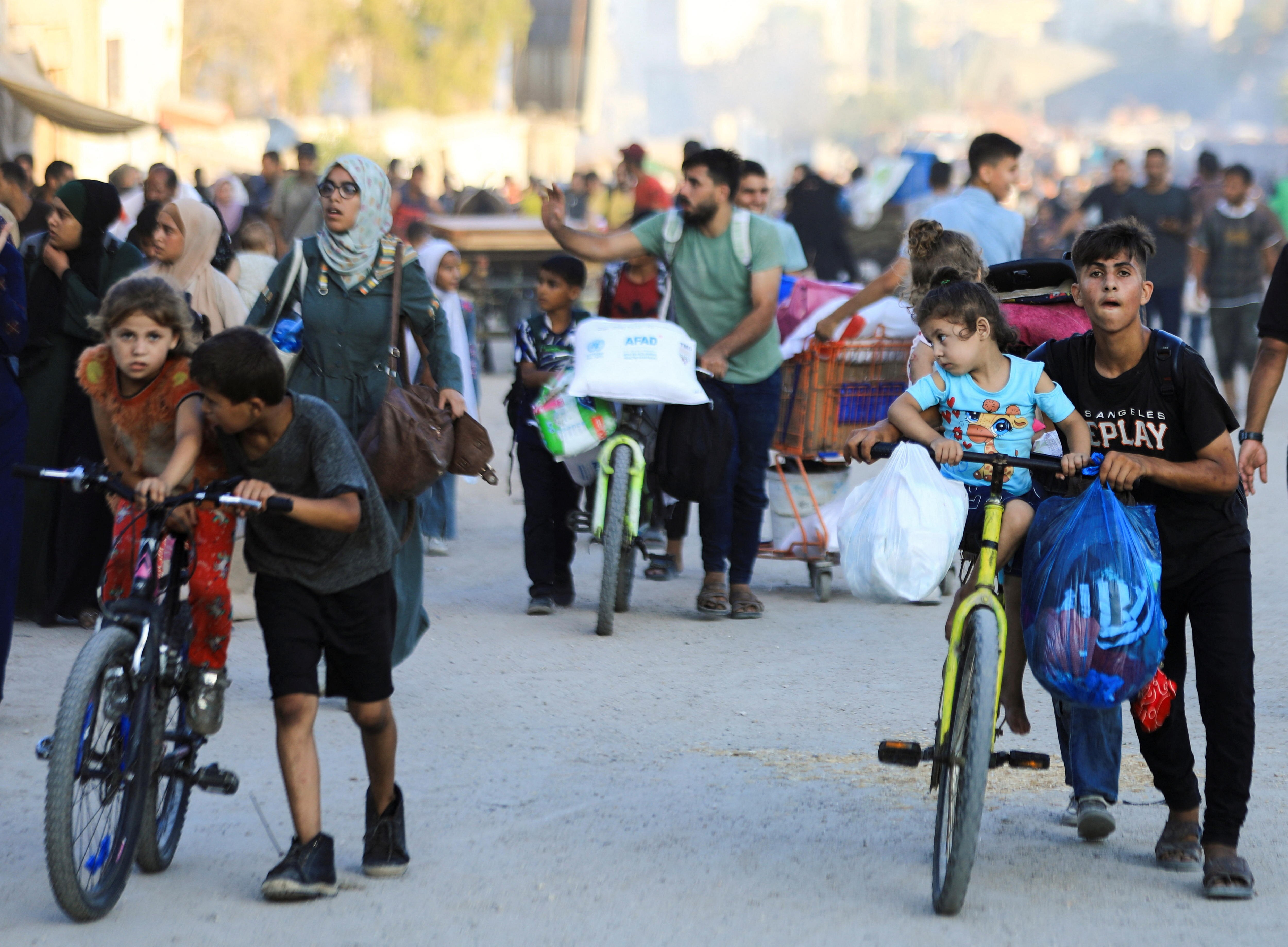 People walk carrying plastic bags and backpacks, two young boys push their bikes with two little girls sitting on the seat