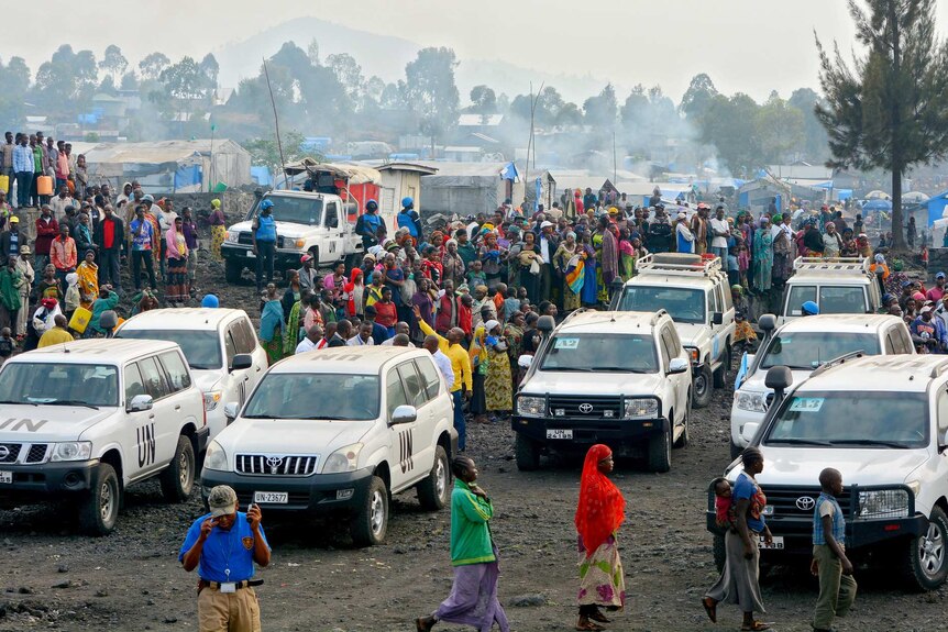 An aerial photo shows a swarm of people in a refugee camp around a number of stationary white UN four-wheel-drive vehicles.