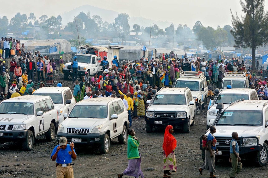 An aerial photo shows a swarm of people in a refugee camp around a number of stationary white UN four-wheel-drive vehicles.
