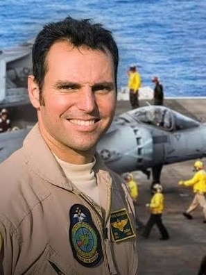 Man in airforce uniform with planes on a ship behind him. 
