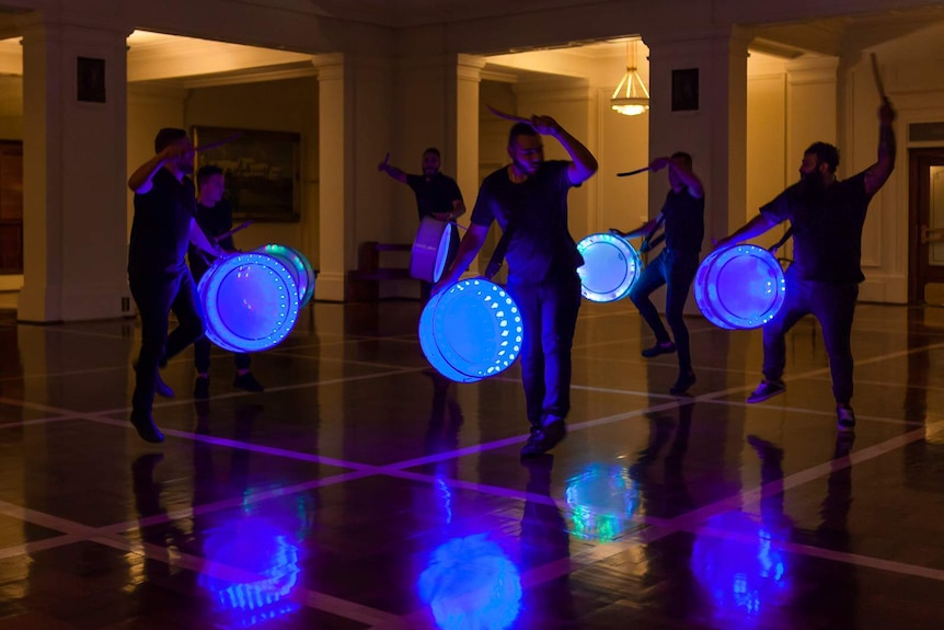 Group of men performing drumming on blue-neon-lit drums, in a darkly lit chamber featuring pillars.