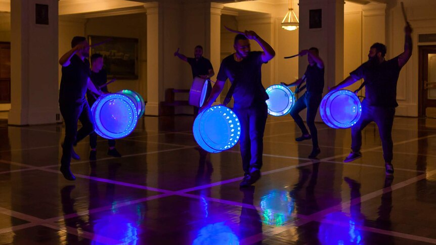 Group of men performing drumming on blue-neon-lit drums, in a darkly lit chamber featuring pillars.