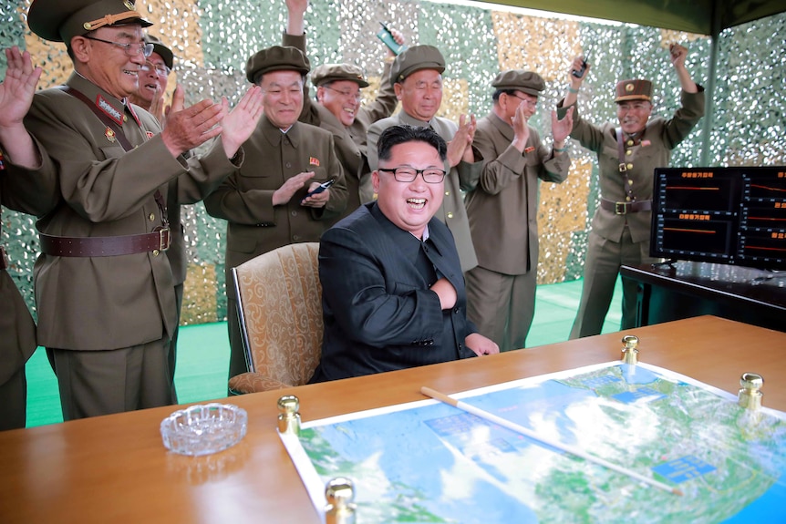 Kim Jong Un with a big smile on his face while men in military dress uniforms stand behind him applauding