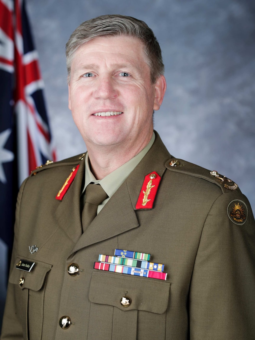 A man stands in military uniform smiling in front of an Australian flag.