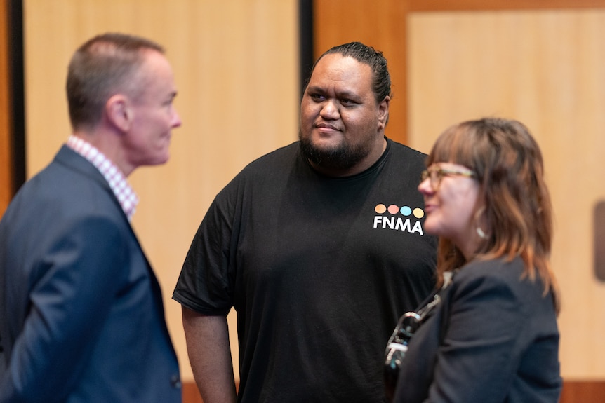 A man is pictured wearing a black t-shirt with the letters 'FNMA' and a logo. He is talking to two people who are out of focus.