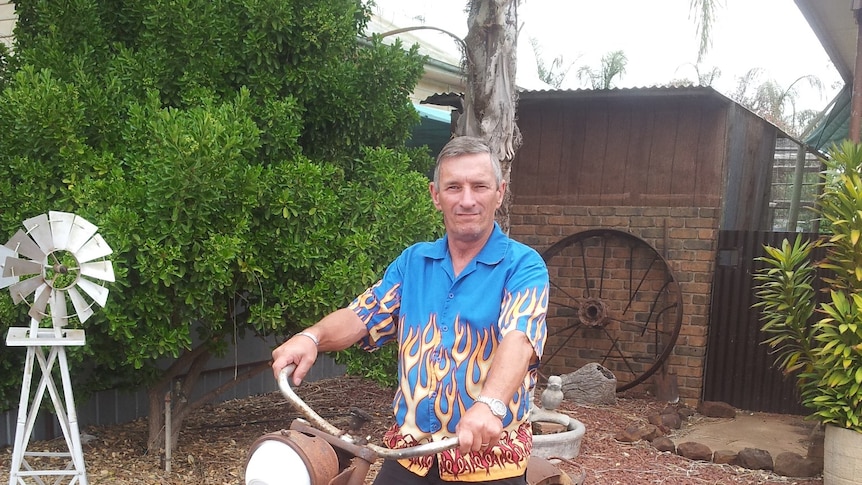 Noel Payne stands astride a rusty old motorbike in a backyard, holding the swept back handlebars, wearing a shirt with flames on