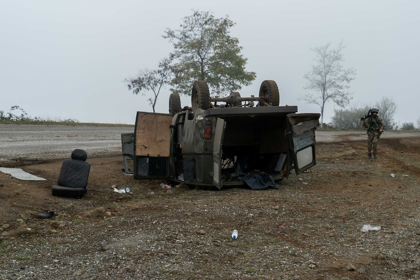 A truck lies on its side next to the road and a soldier walks past