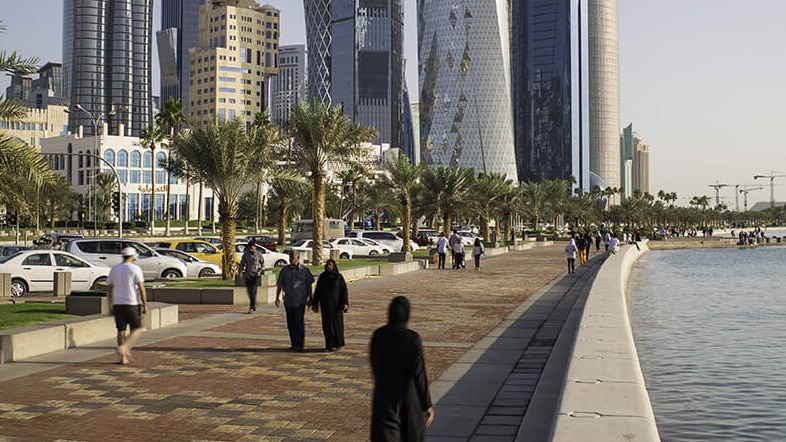 Huge towers dominate the Doha CBD skyline while people walk along a footpath.