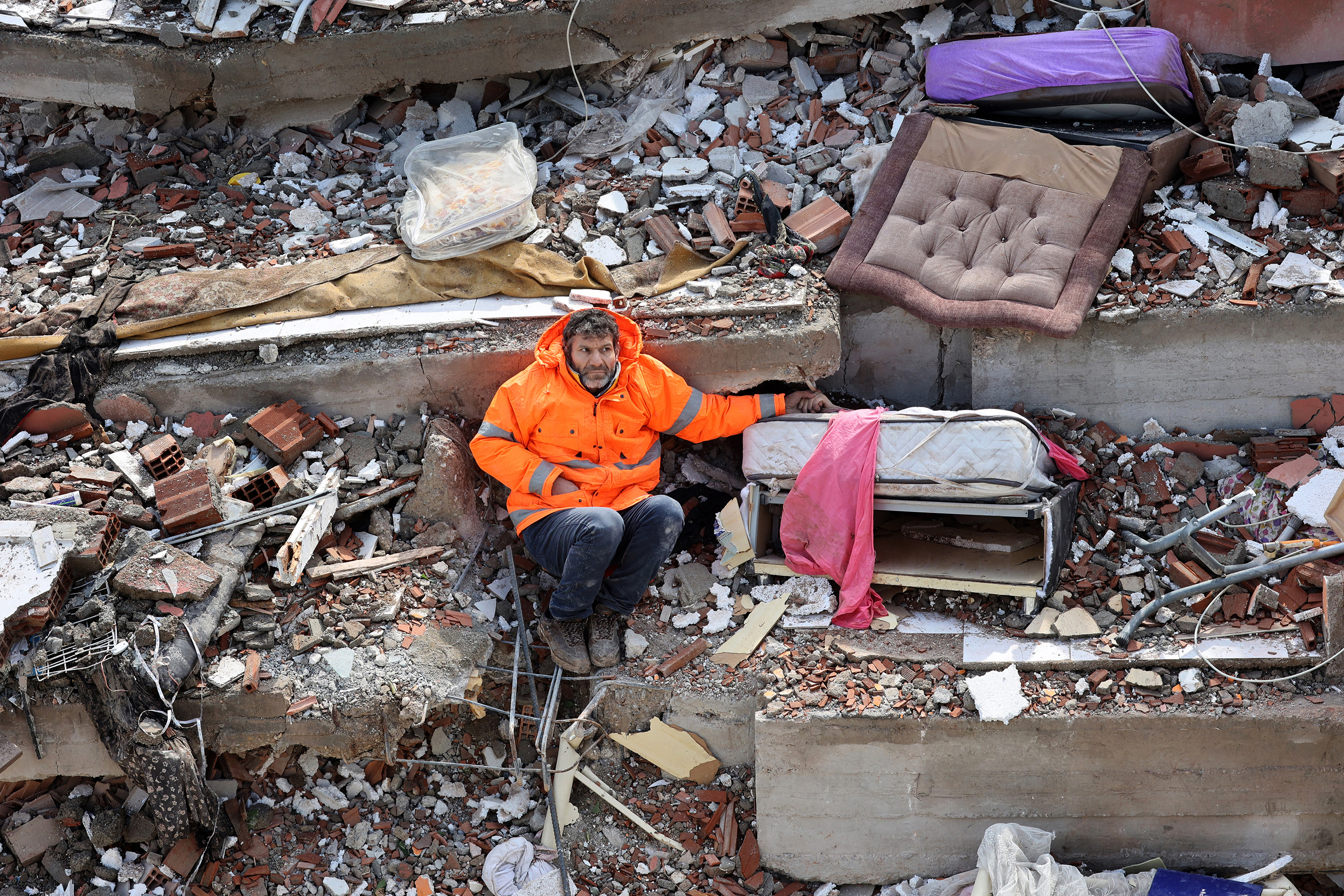 A man in an orange high-visibility jacket sits among the rubble of a building destroyed by a earthquake