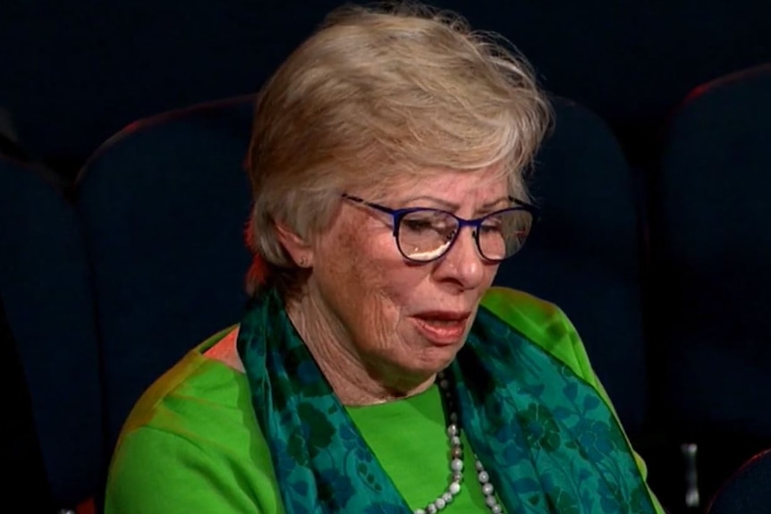 A woman with short light hair and glasses wears a bright coloured top and scarf.