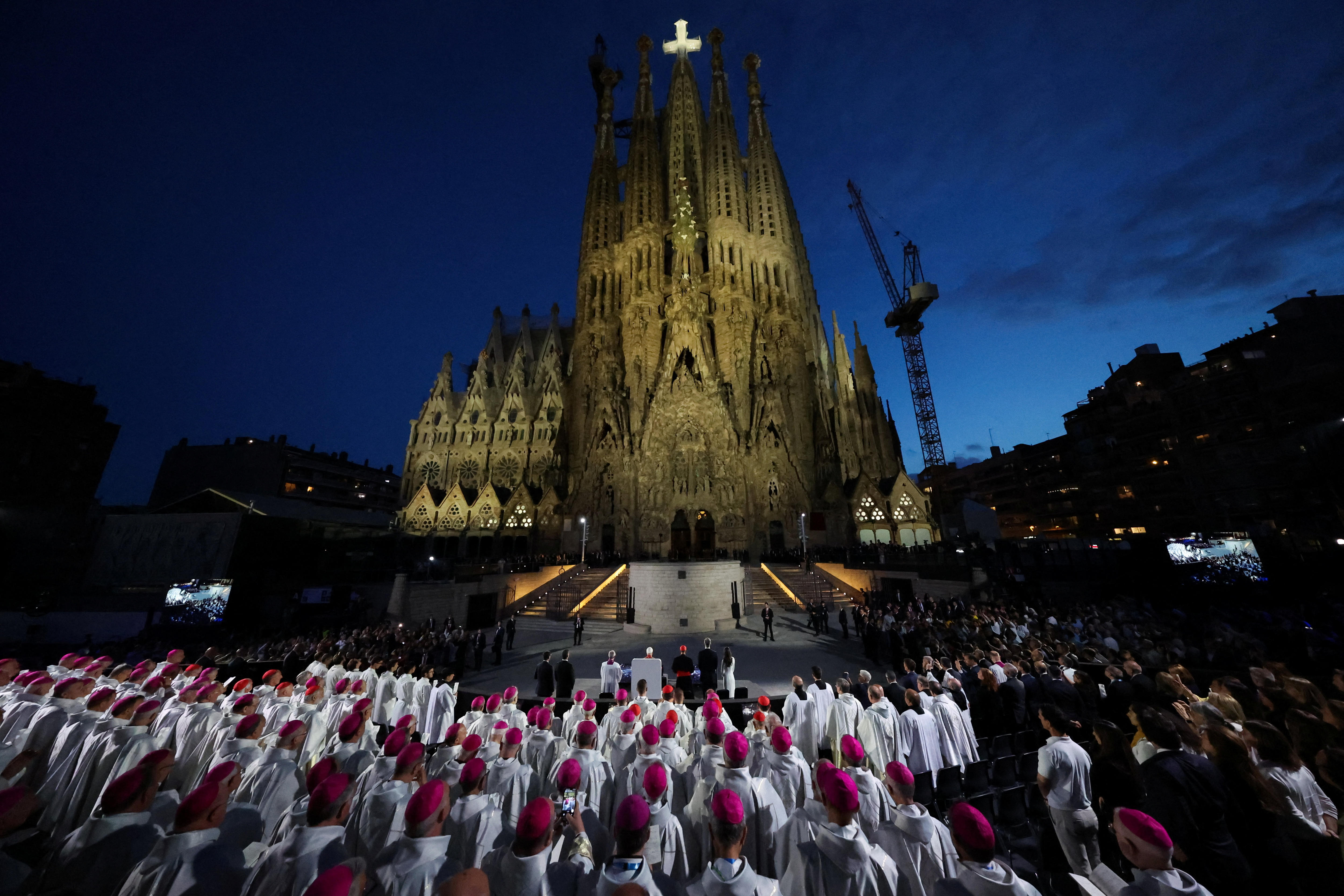 The Sagrada Familia at night, illuminted in light with a white cross at its top, surrounded by people in white robes.