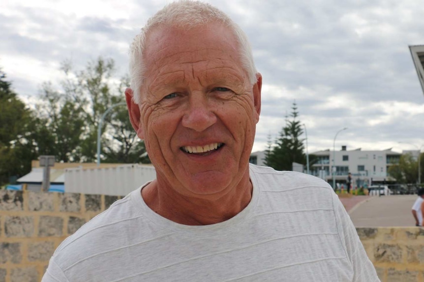 An older man with a white shirt smiling at the camera on the beach front