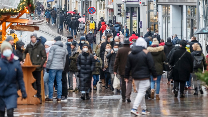 People walk down a street, some with umbrellas, most wearing masks