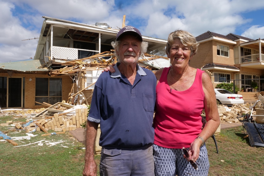 An older man in a blue shirt and a woman in a pick shirt stand in front of a house destroyed by a cyclone.