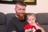 A medium shot of Nicholas wearing a dark grey t-shirt, holding his toddler son James, sitting on a couch, smiling at the camera.