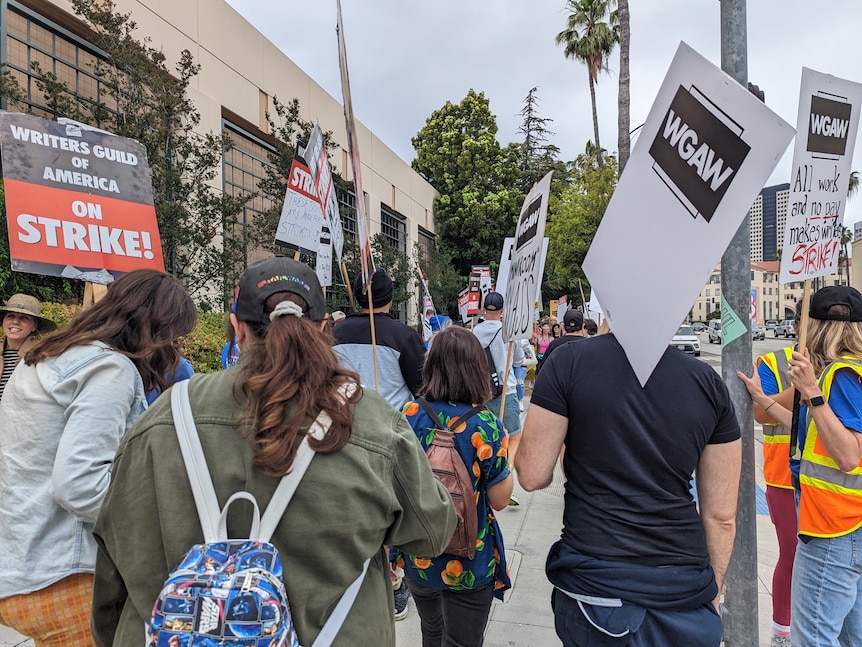 People visible from the back holding signs and walking while on strike