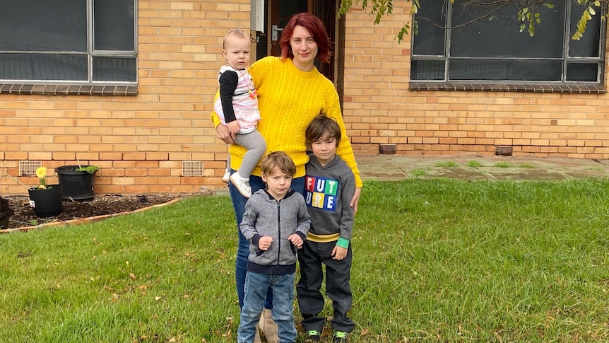 A woman wearing a yellow jumper with her three young children outside a brick home.