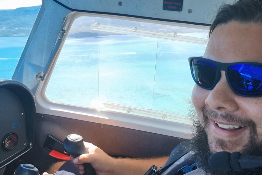 a man smiling at the controls of a small plane over a tropical sea