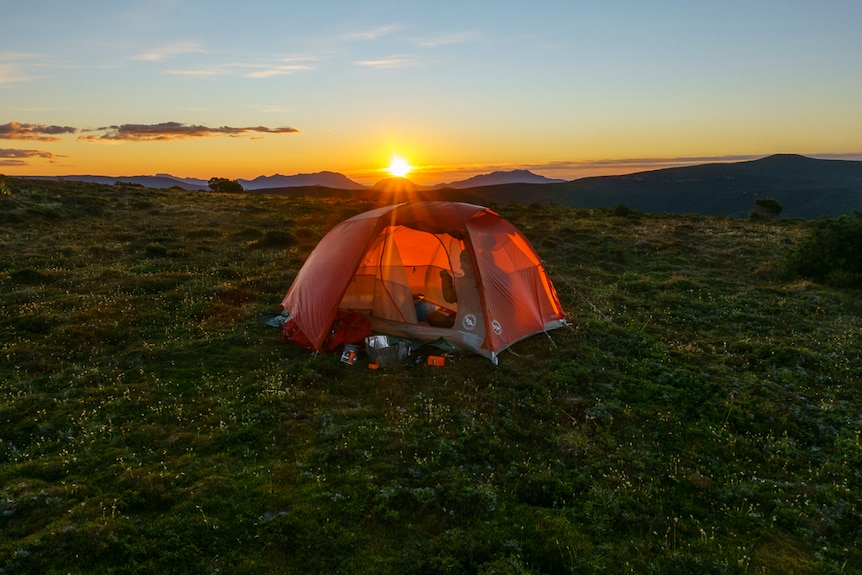 an orange tent up high on a mountain, the sun is setting behind it