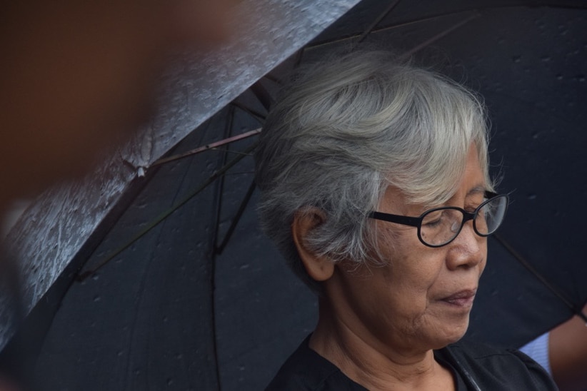 Close up picture of a woman with short white hair, wearing glasses, under a black umbrella.