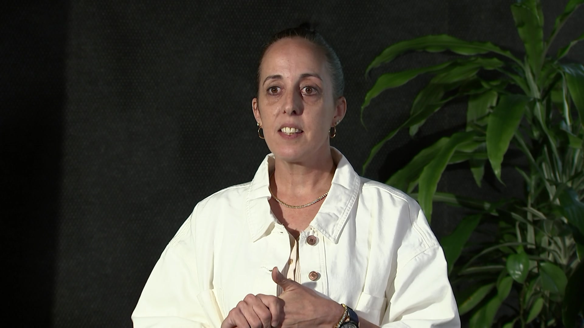 Woman in white jacket looks at an interviewer to the left of frame. There is a black backdrop behind her + a green plant.