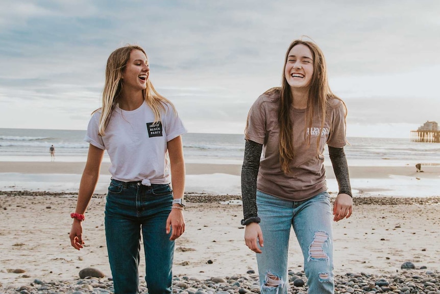 Two young women laugh as they walk together on a pebbled beach.
