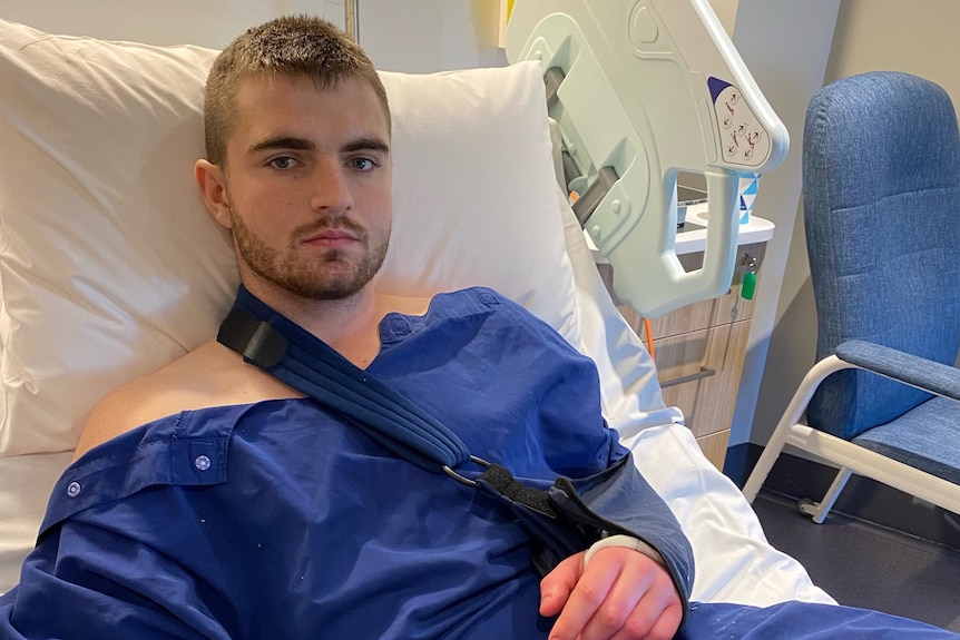 A young man in blue hospital scrubs in a hospital bed with a sling around his shoulder. He looks tired.