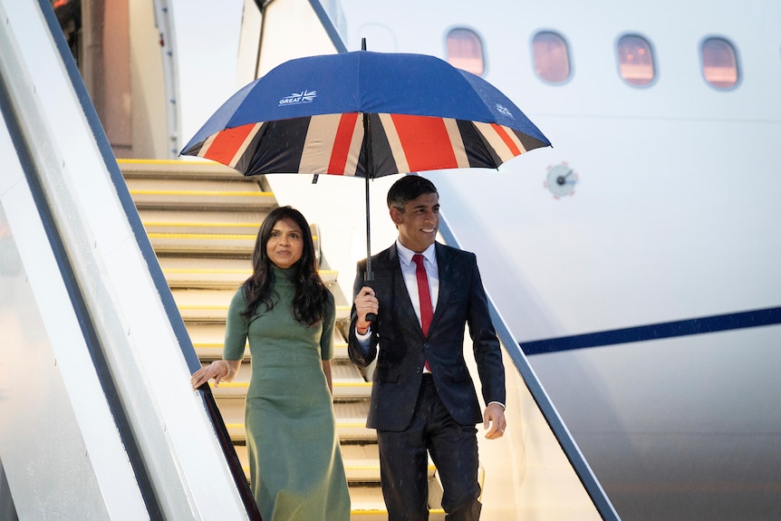 Rishi Sunak and his wife walk down the steps of a plane holding a union jack flag umbrella