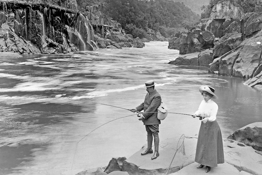 A black and white photo of a man and woman in old-fashioned clothing fishing by a river