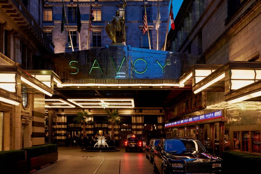 The exterior entrance of the Savoy Hotel at night. Flash vintage cars are parked out the front.