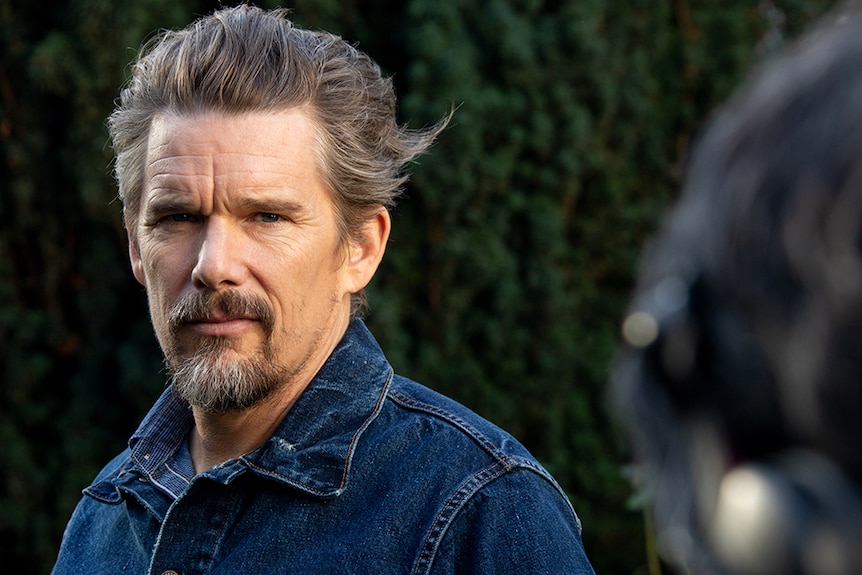 A man with short greying brown and facial hair wears dark denim jacket and stands outdoors in front of trees.