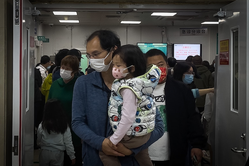 A man carrying a child walks out from a crowded holding room with children.