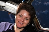 Anne McClain sits at an angle, holding onto a bar, in a pod on the International Space Station