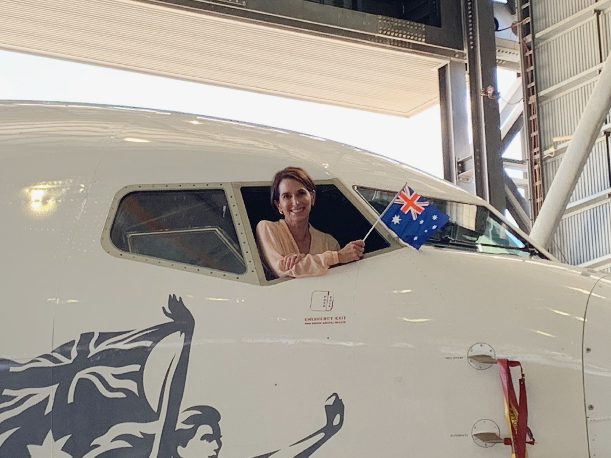 Virgin CEO Jayne Hrdlicka sits in the cockpit of a plane, waving an Australian flag.