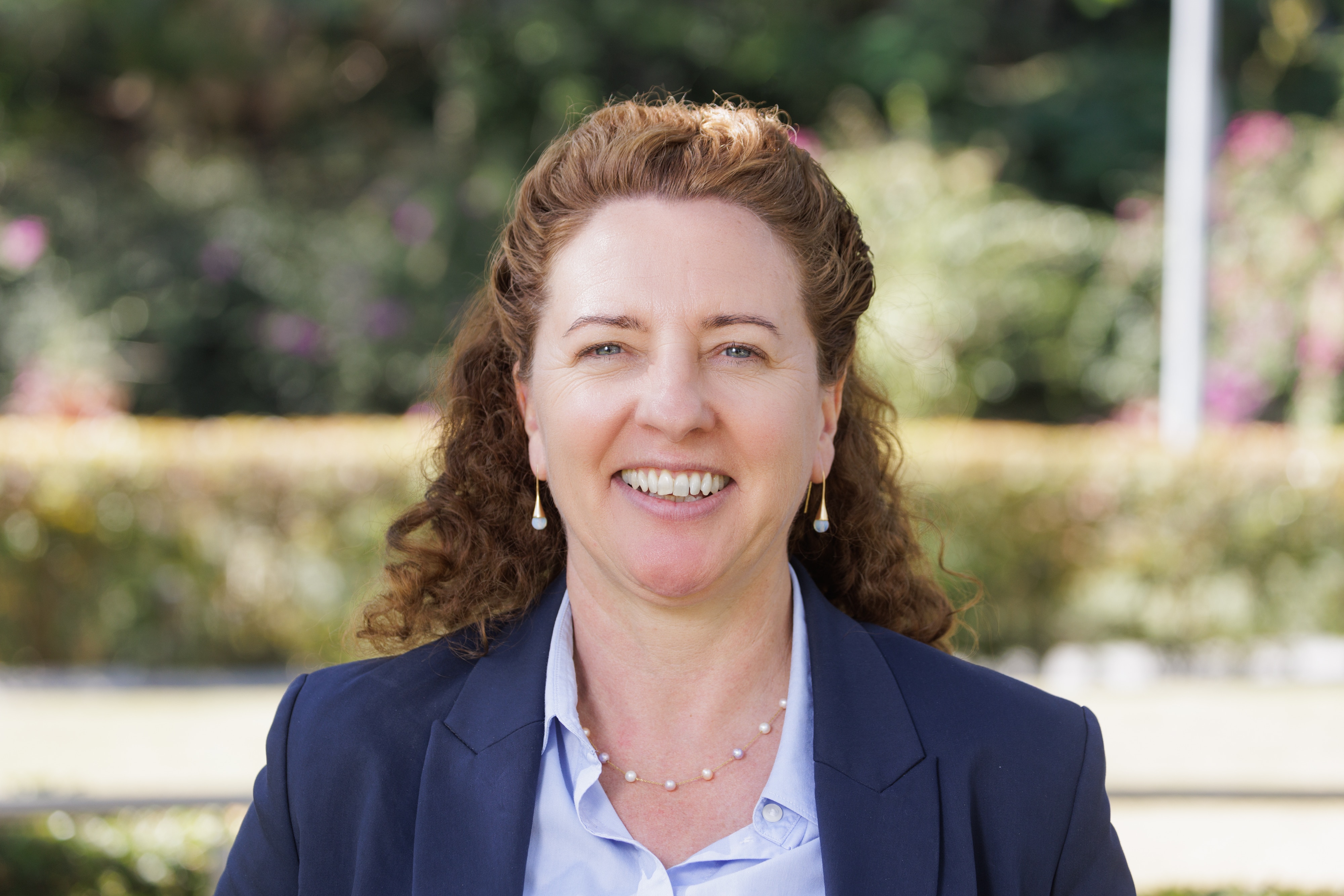 A headshot of a smiling woman with red-brown hair, wearing a light blue shirt and navy blazer.