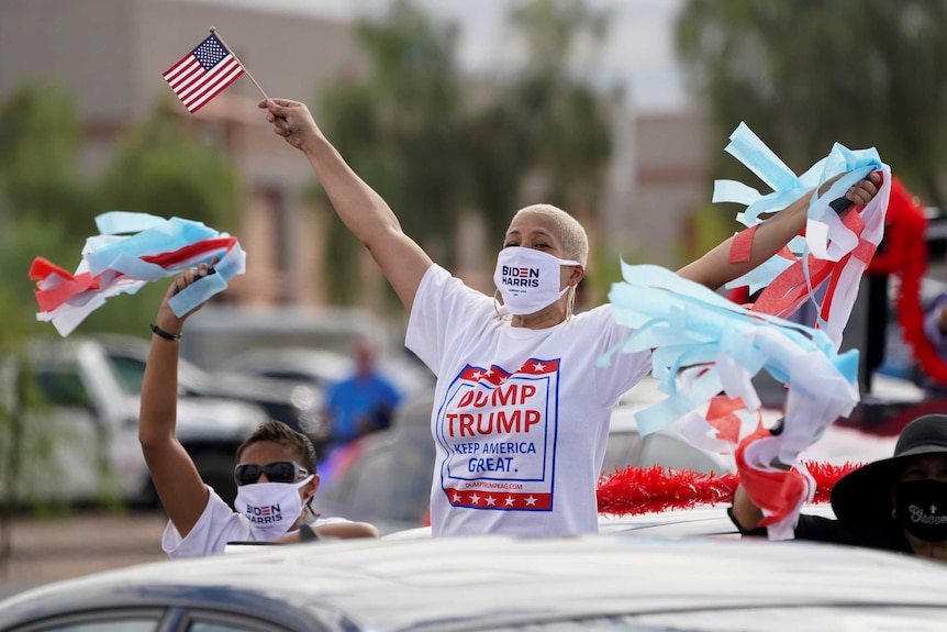 A woman wearing a 'dump Trump' t shirt and Biden/Harris mask waves streamers and an american flag at a Joe Biden campaign rally