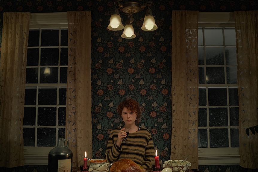 A woman with red curly hair with serious expression holds wine glass and sits at dinner table with food in stylised interior.